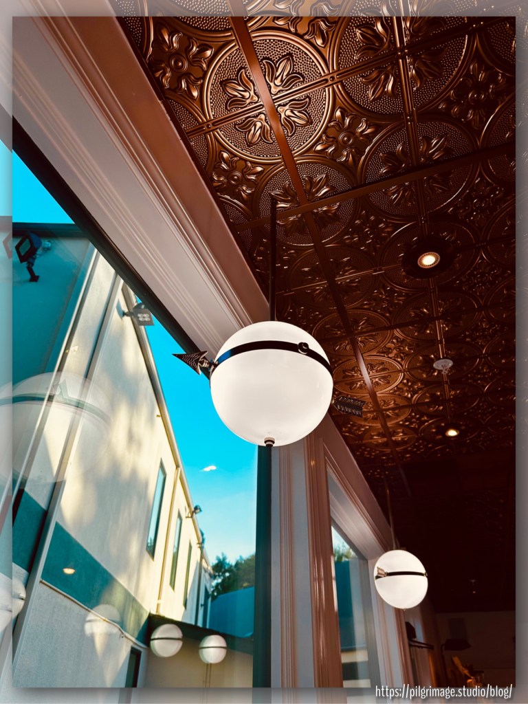 Gilded ceiling panels, the globe lights glow with an arrow protruding from their centers overlooking a bright blue sky outside the window. Globe light reflections hang suspended in the glass.